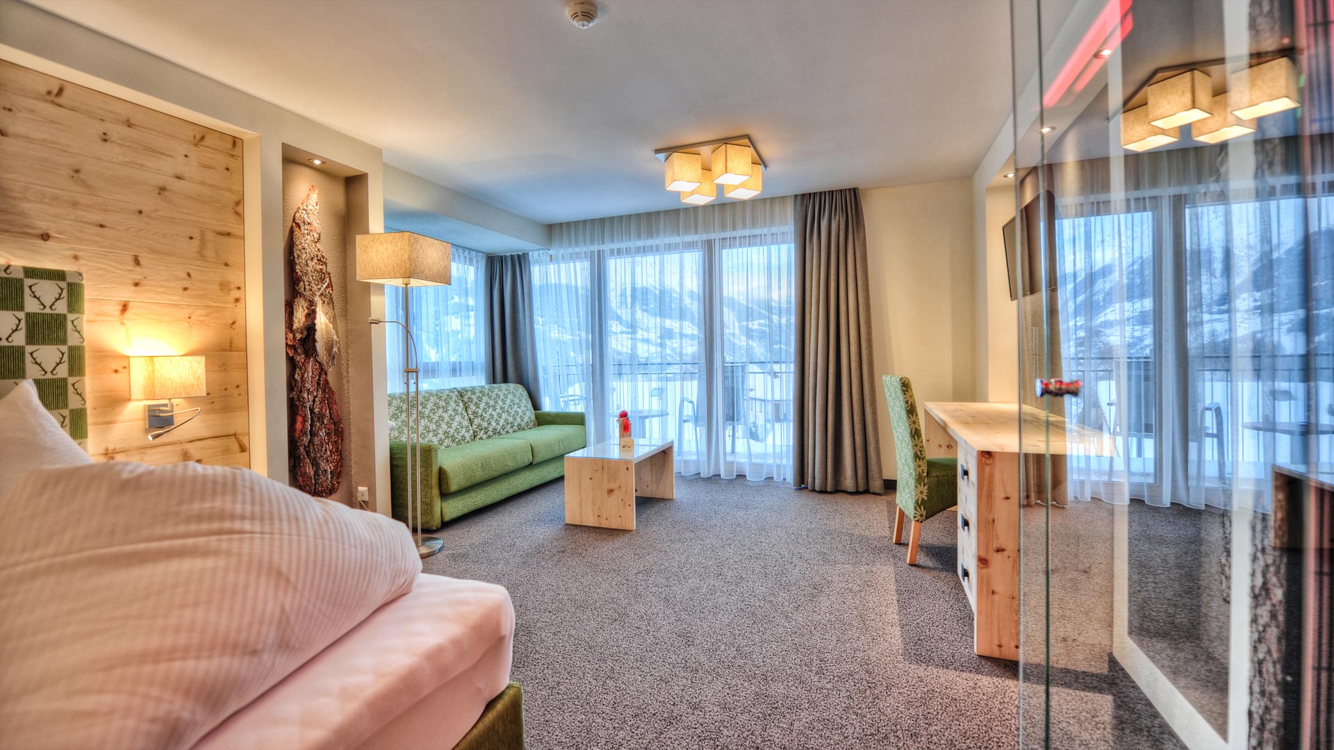 Helles Hotelzimmer mit Bergblick, grünem Sofa, Schreibtisch, Doppelbett, Holzmöbeln und großen Fenstern mit Vorhängen.